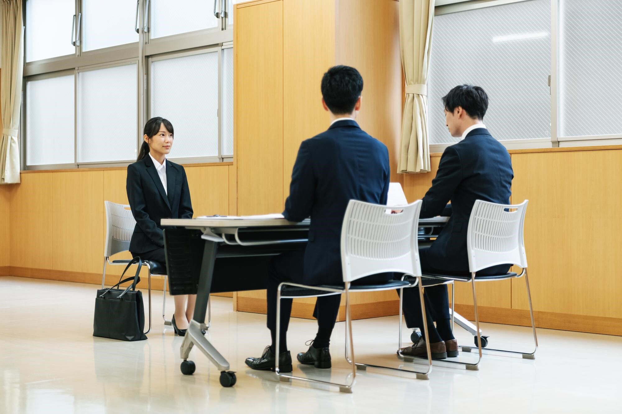椅子に座るスーツ姿の女性と机を挟んで向かいに座る男性2人の後ろ姿