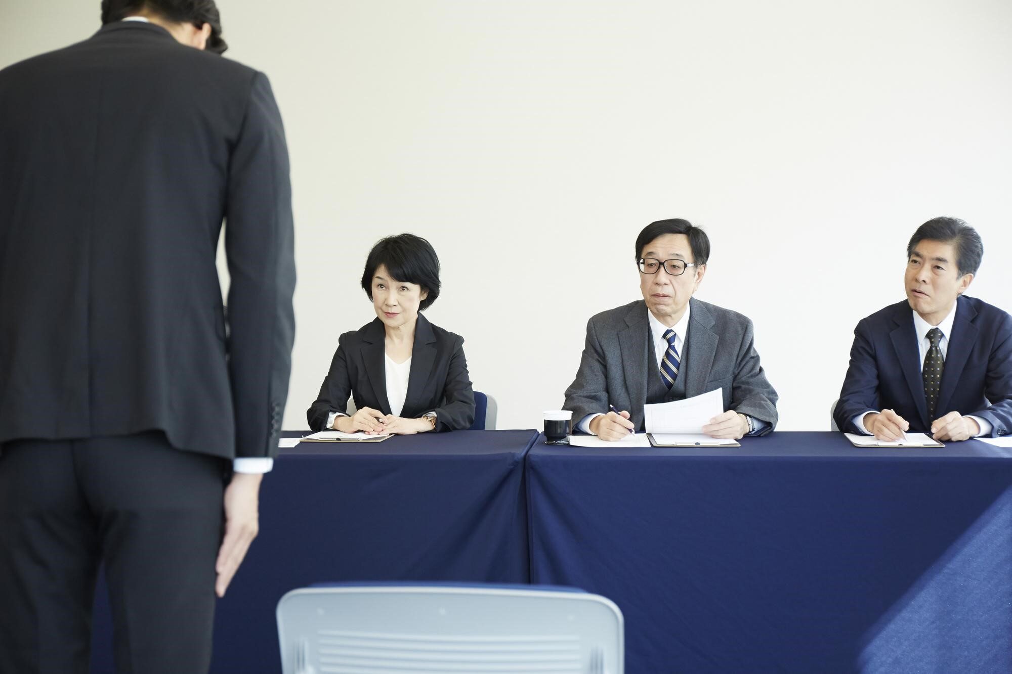 お辞儀をするスーツ姿の人物の後ろ姿と正面の机に向かって座る3人の男女